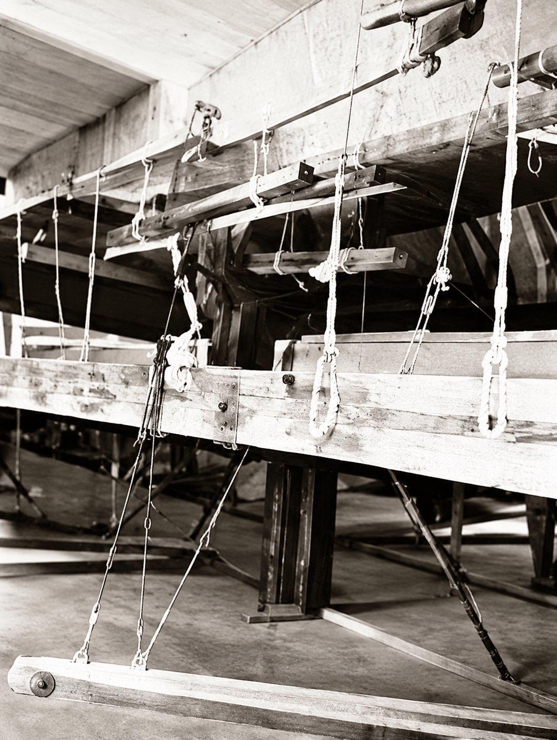 Photo en noir et blanc de l'intérieur d'un atelier où des plateformes en bois sont suspendues par des cordes.