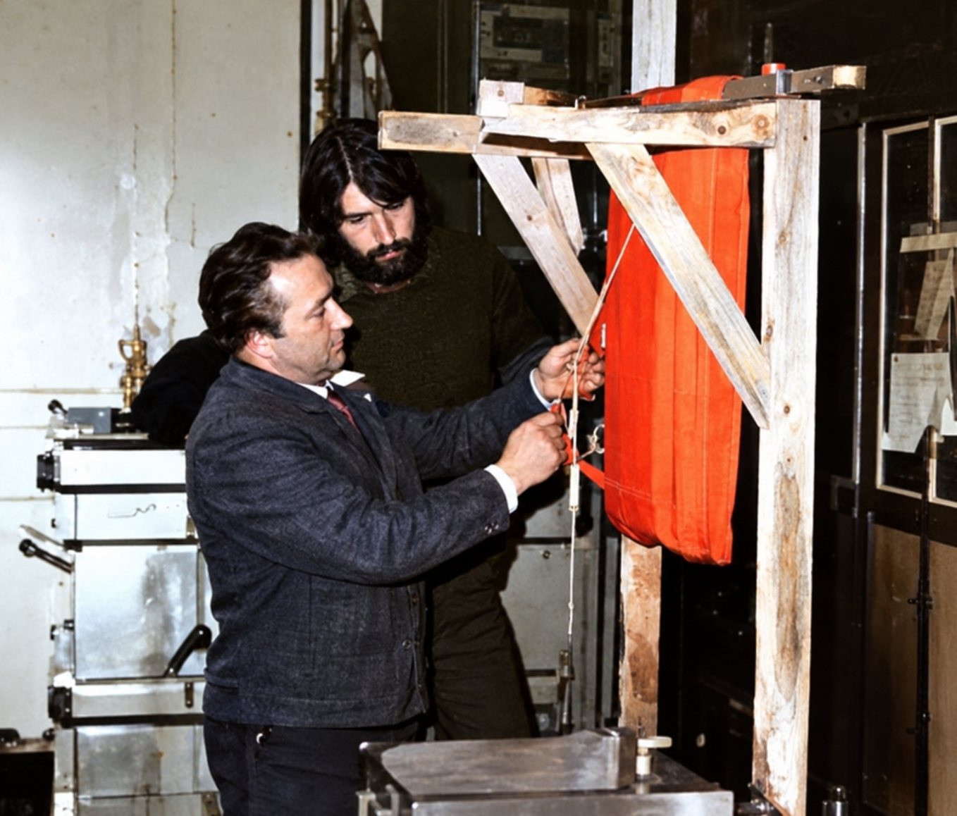 Deux hommes ajustent une structure en bois sur laquelle est suspendu un grand tissu orange, dans un atelier au mur clair.