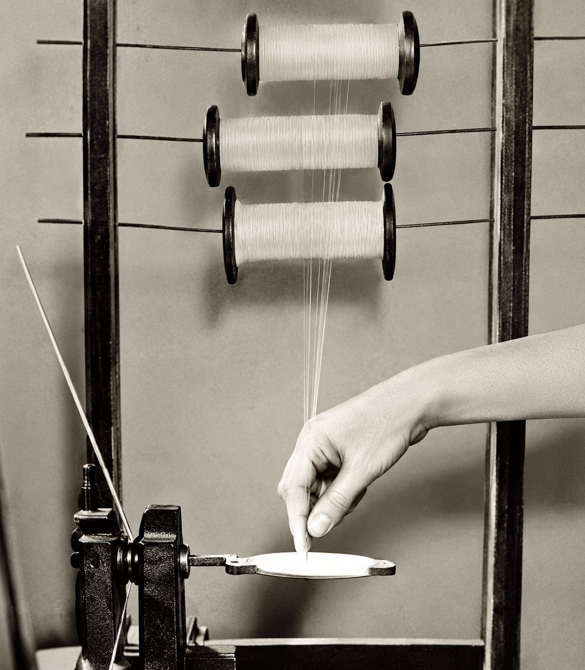 Photographie en noir et blanc d'une main qui tient un fil lumineux devant plusieurs bobines montées sur une machine textile.