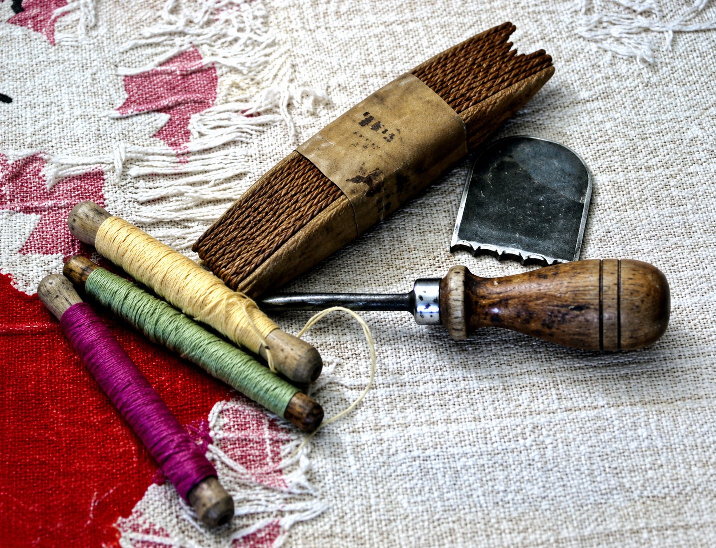 Une alène à manche en bois, des bobines de fil colorées et un petit outil en bois posés sur un tissu brodé à motifs clairs.